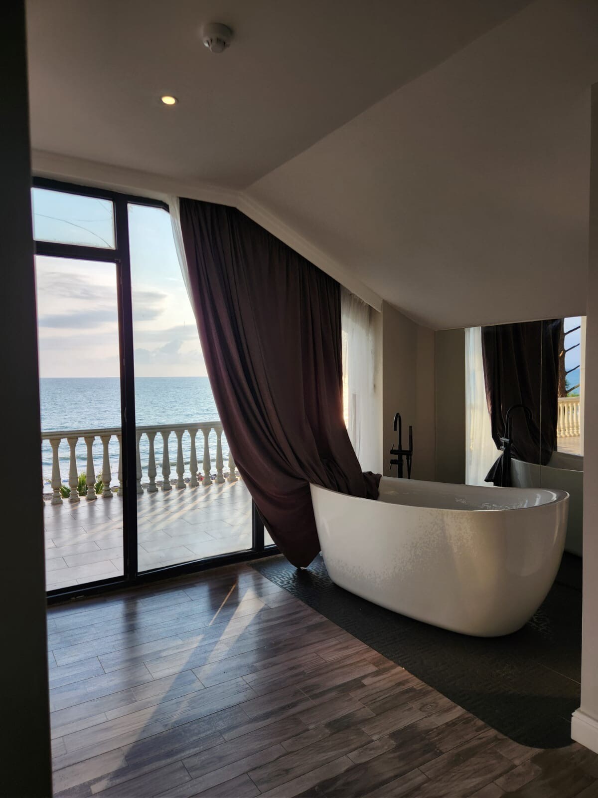 A spacious bathroom features a modern freestanding tub positioned near large glass doors that provide a view of the sea. Floor-to-ceiling windows are dressed with flowing curtains, and a stylish vanity is reflected in the adjacent mirror, enhancing the overall elegance.