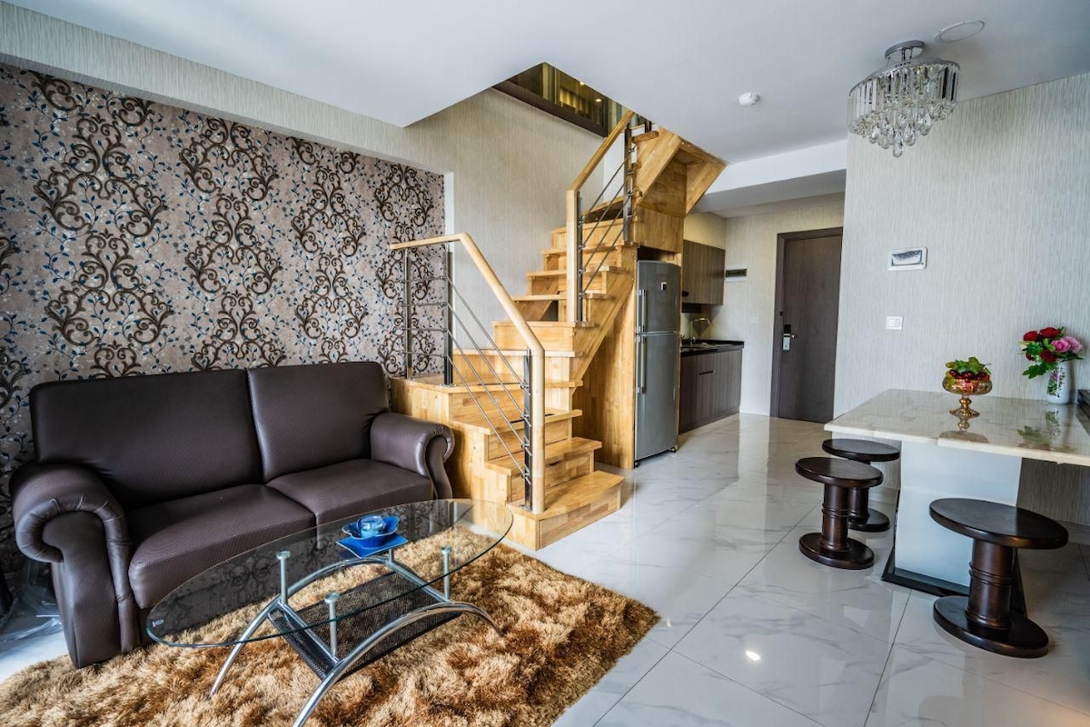 A stylish living area is featured, showcasing a dark brown sofa paired with a glass coffee table on a plush rug. A wooden staircase leads to the upper level, with an adjacent dining table surrounded by two matching stools. The backdrop includes a decorative wall with intricate patterns.