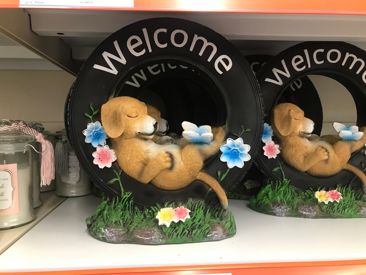 A unique welcome sign features a dog lounging inside a decorative tire. The dog is depicted in a relaxed position, surrounded by colorful flowers and greenery. The word 'Welcome' is displayed prominently at the top of the tire.