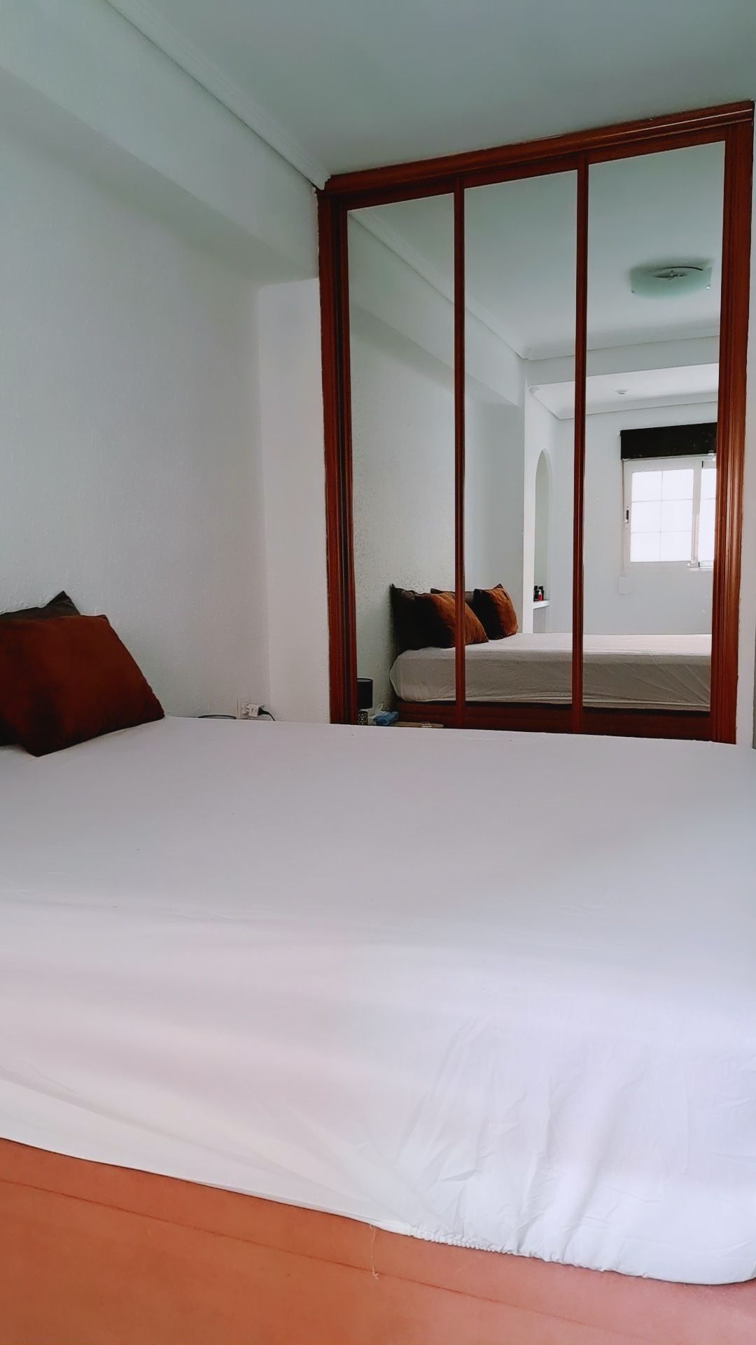 A spacious bed is neatly dressed in white linens, with upholstered pillows placed at the head. A large mirrored wardrobe reflects the room's light and creates a sense of openness. Natural light filters through the window, enhancing the calm atmosphere.