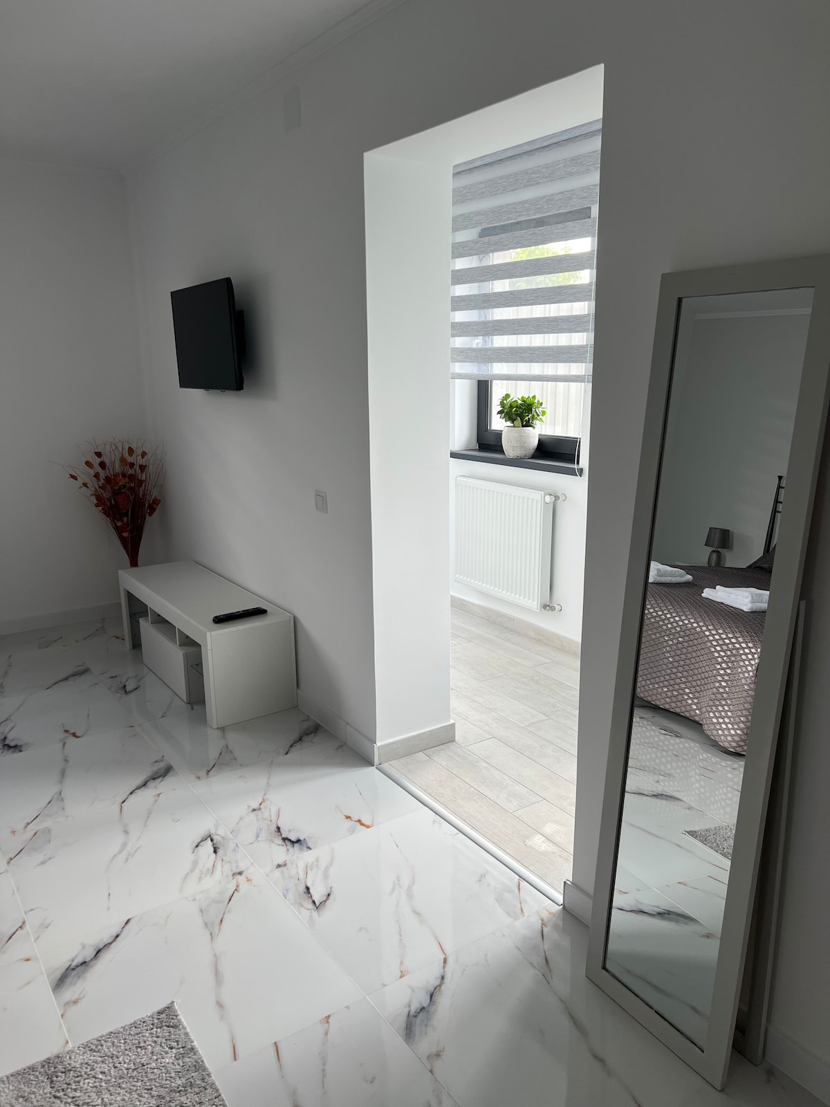 A bright and airy room is highlighted by sleek marble flooring. A large mirror reflects the space, while a wall-mounted television adds a modern touch. Natural light filters through layered window treatments, illuminating a glimpse of a cozy bedroom through the open doorway.