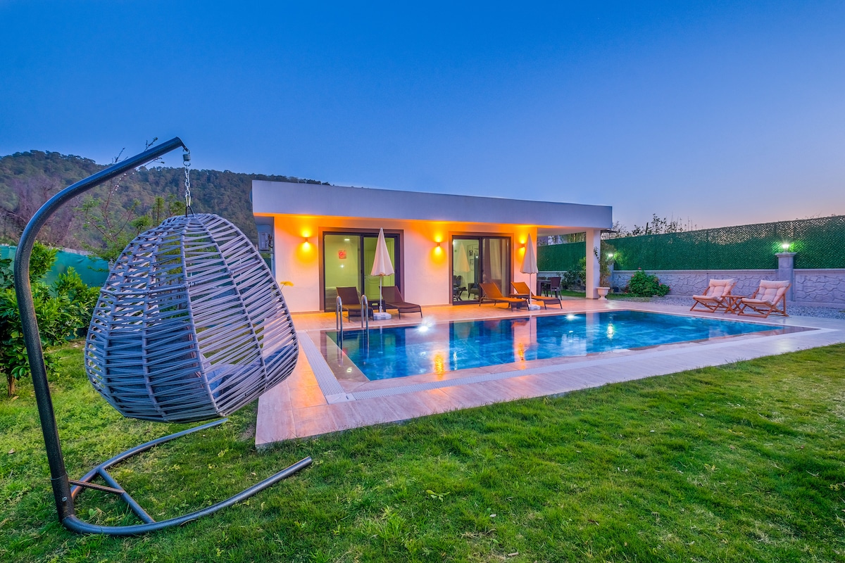 The outdoor area features a private swimming pool surrounded by lush greenery. A hanging chair is positioned beside the pool, alongside lounge chairs and a sun umbrella. Warm lighting highlights the villa's exterior, creating an inviting setting during dusk.