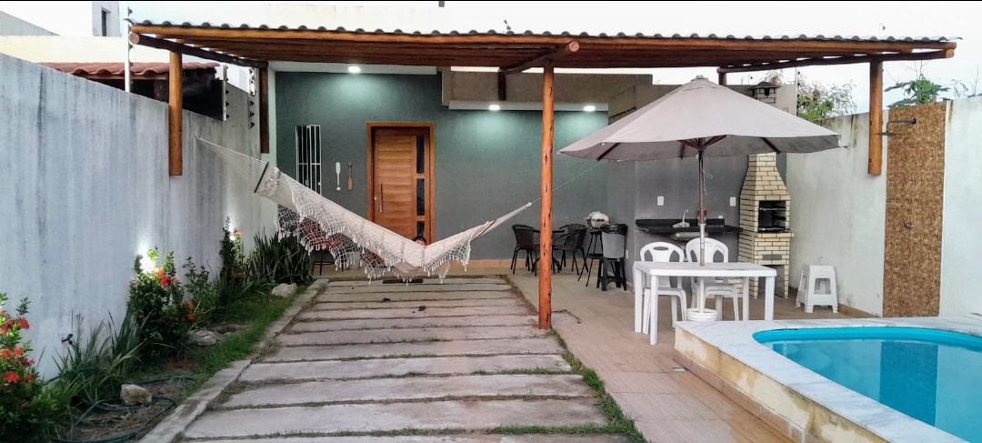 An outdoor patio features a shaded seating area with a table and chairs. A hammock hangs from the wooden structure, while a grill is positioned nearby. Lush greenery and flowers border the stone paths, leading to a pool that reflects the evening light.