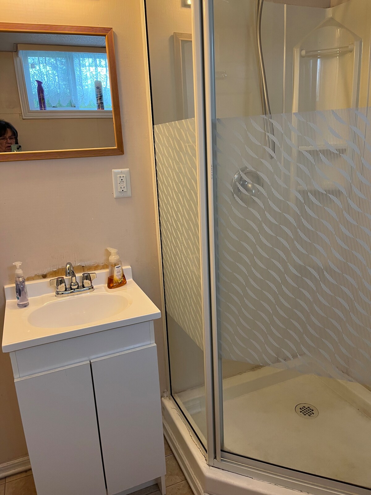 A bright and clean bathroom is shown, featuring a compact white sink cabinet with faucet and toiletries. A glass shower enclosure with frosted wave patterns is visible, highlighting the showerhead and curved walls. Natural light enters through a nearby window, enhancing the inviting atmosphere.