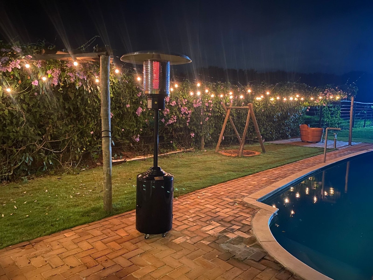 An outdoor area is illuminated by string lights, casting a warm glow over the space. A tall heater is positioned nearby, providing warmth. A swimming pool reflects the lights while a grassy area adds to the environment. The setting enhances evening gatherings.