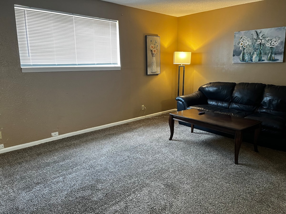 The living room features a large window allowing natural light to fill the space. A comfortable black couch is positioned alongside a wooden coffee table. Soft carpet covers the floor, and a floor lamp adds additional lighting. Two pieces of wall art are displayed on the light brown walls.