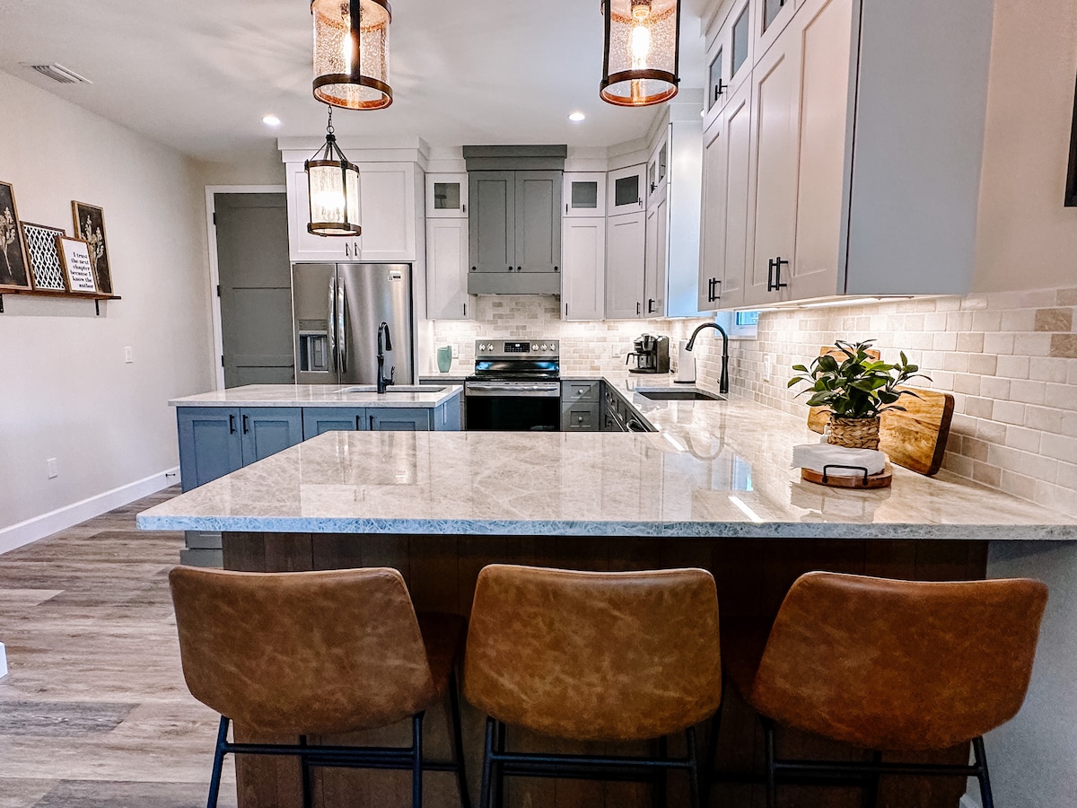 A modern kitchen features light gray cabinets and a large island with seating for three. The countertops are made of a light-colored stone, and stainless steel appliances are visible. Warm lighting from hanging fixtures adds a welcoming touch to the space.
