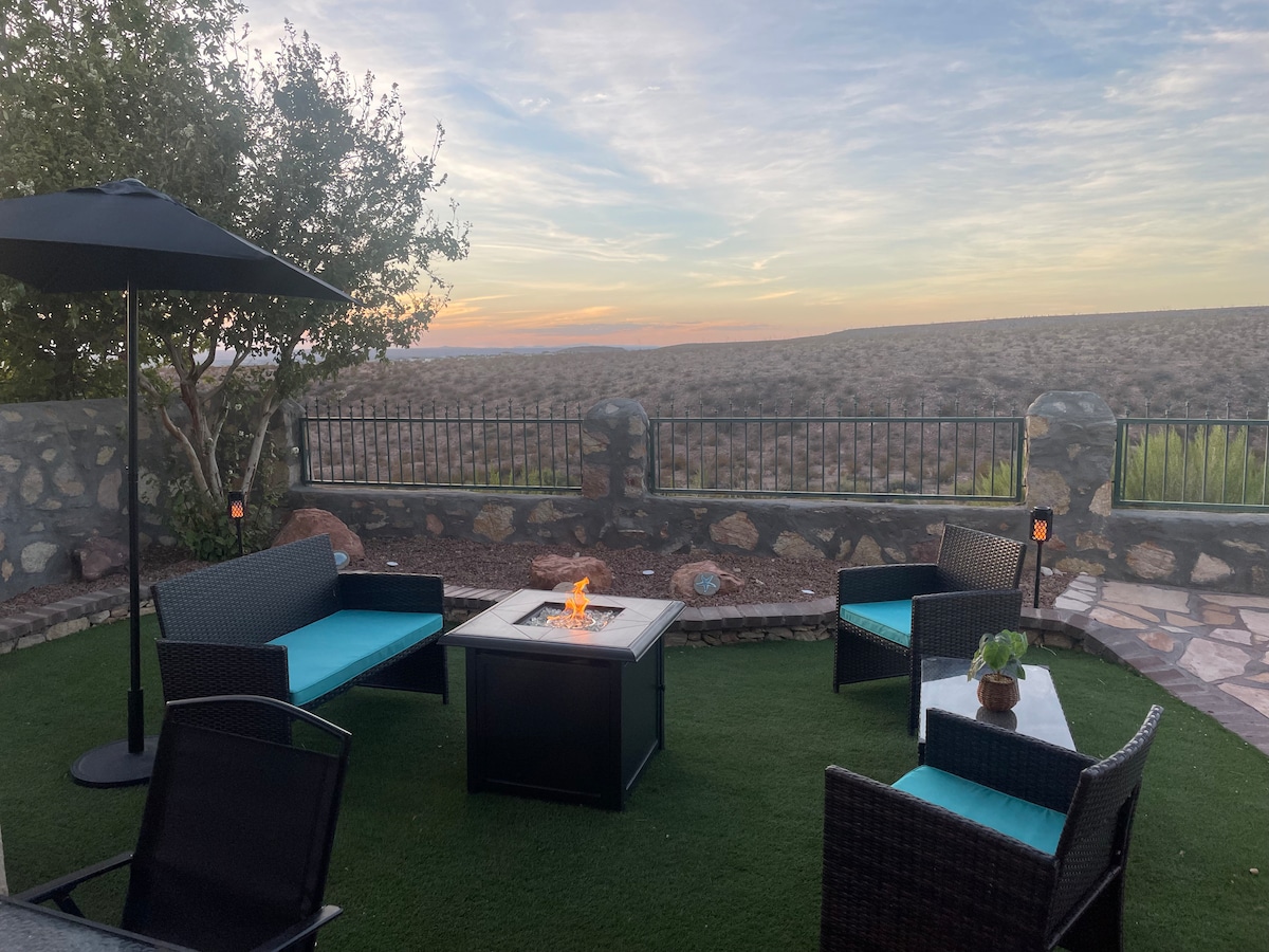 An outdoor seating area features modern black wicker furniture with teal cushions, arranged around a central fire pit. Surrounding stones and greenery enhance the space, with a vast landscape visible in the background under a soft sky.