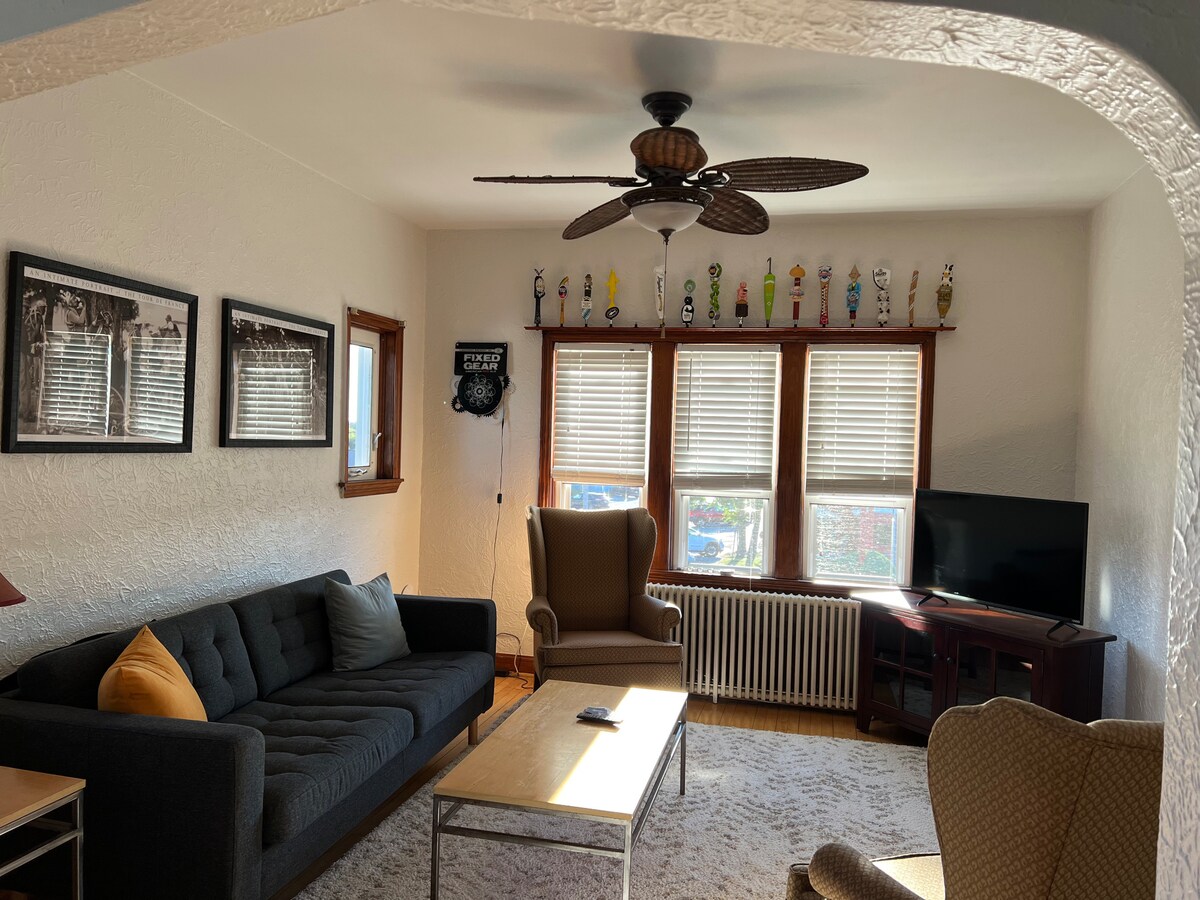 An inviting living area features a dark gray sectional sofa paired with a beige armchair. Natural light filters in through three large windows adorned with blinds. A coffee table sits in the center, complemented by framed photographs on the walls and a television in the corner.