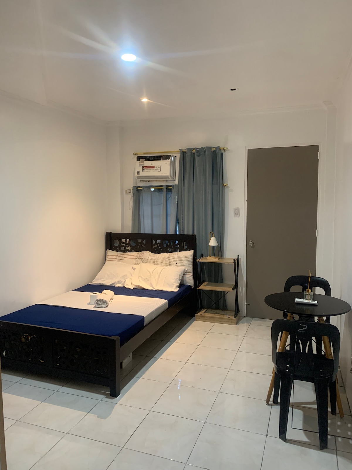 A compact room features a dark wooden bed with white bedding and a decorative headboard. An air conditioning unit is mounted above a window with light curtains. A small round table and two chairs are set against one wall, offering a simple dining option.