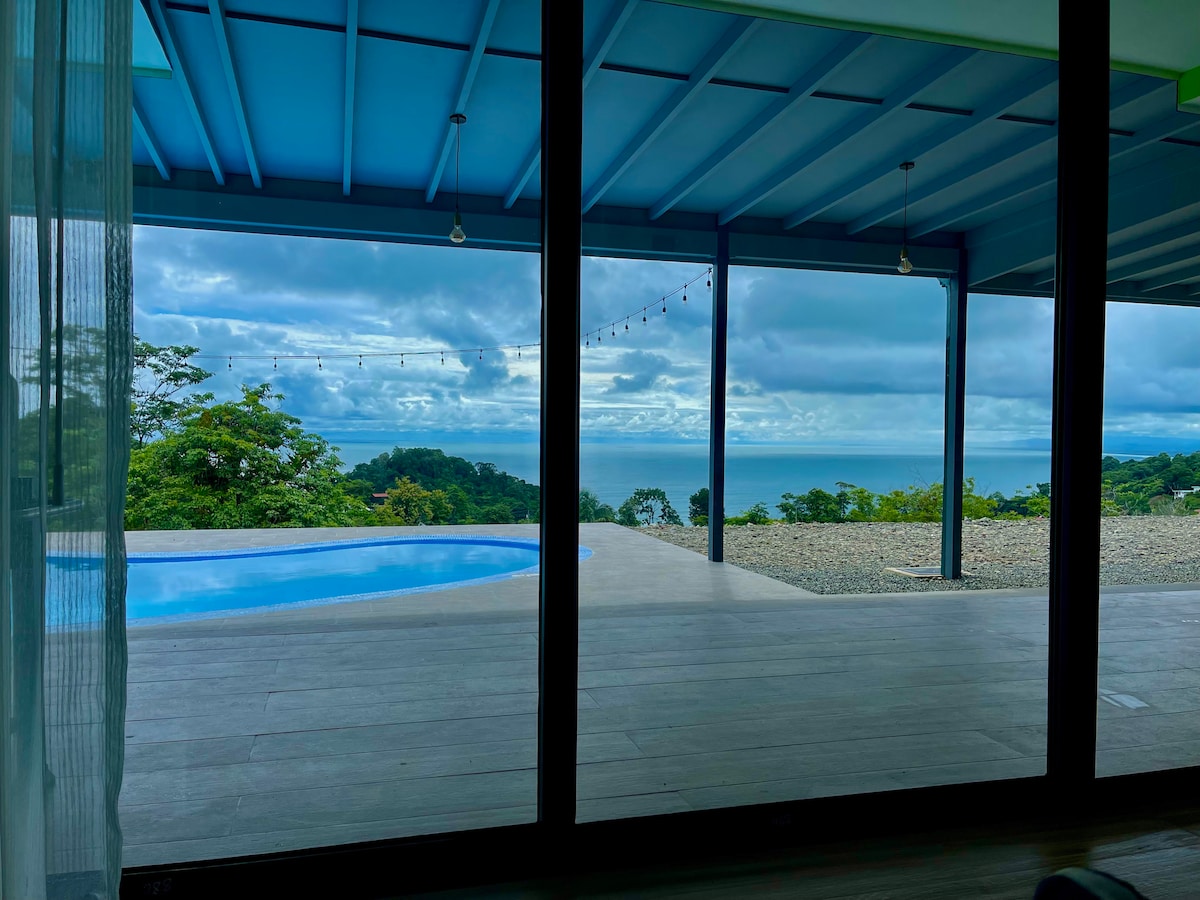 A spacious outdoor terrace features a stunning view of the Pacific Ocean, with a swimming pool reflecting the sky. Glass doors create a seamless transition between indoor and outdoor areas, allowing natural light to brighten the space. Lush greenery surrounds the property.
