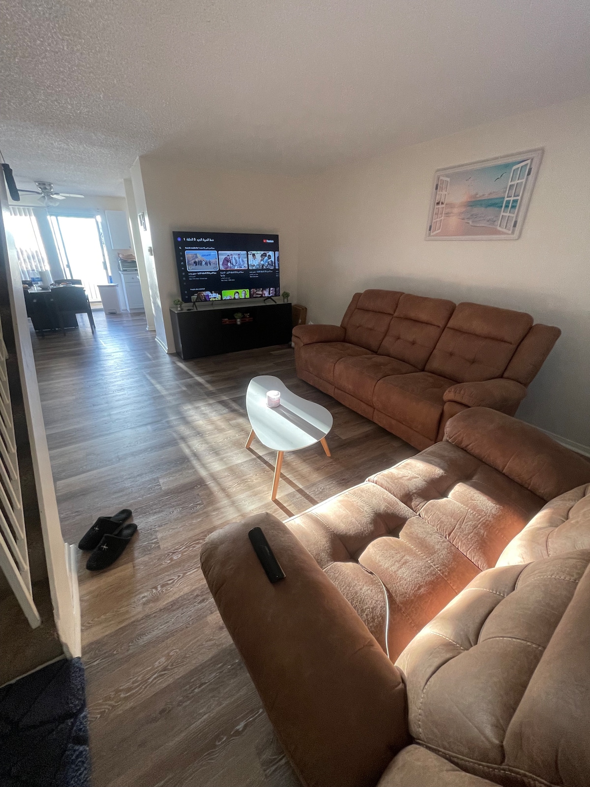 A bright living area is featured with two brown sofas positioned for comfort. A small white side table is placed nearby. A television displays entertainment options, and sunlight is welcomed through large windows. A view of the dining area can be seen in the background.
