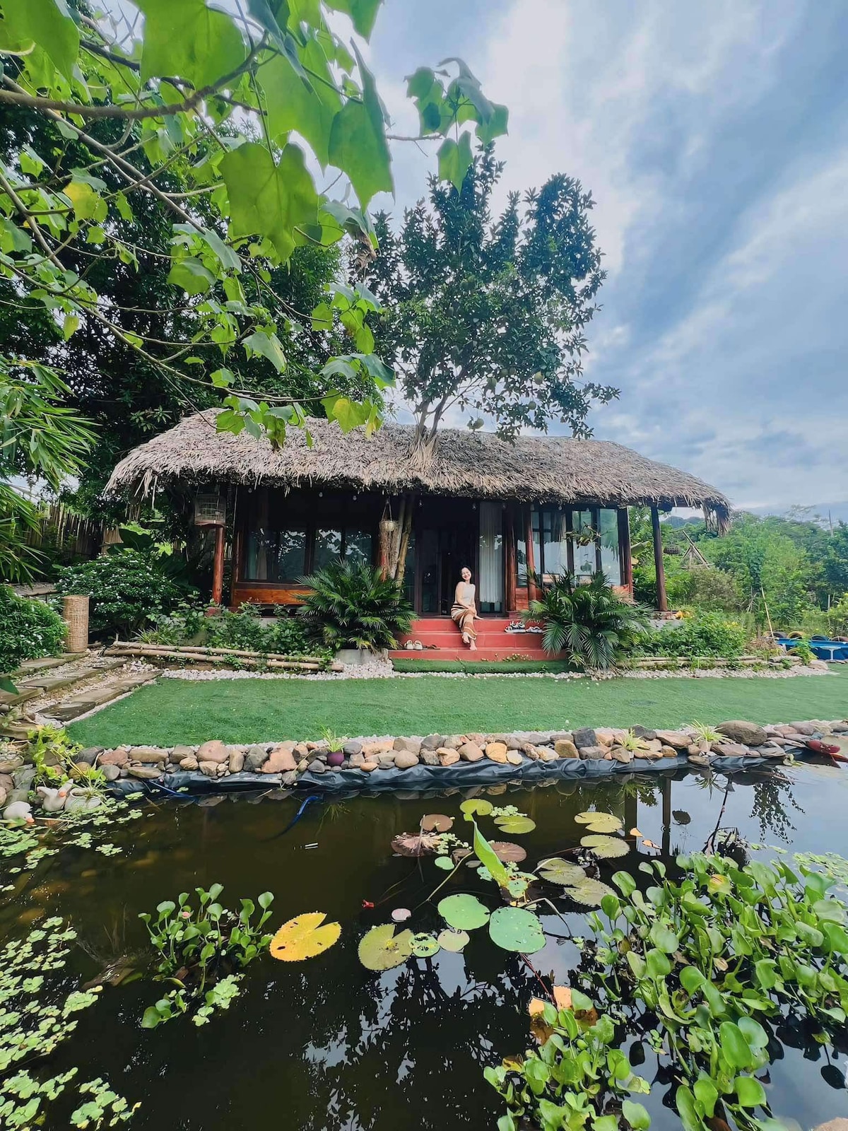 A thatched-roof villa is surrounded by lush greenery and a serene pond. The villa features large windows that invite natural light, with a person standing at the entrance. The pond is dotted with water lilies, complementing the tranquil outdoor setting.