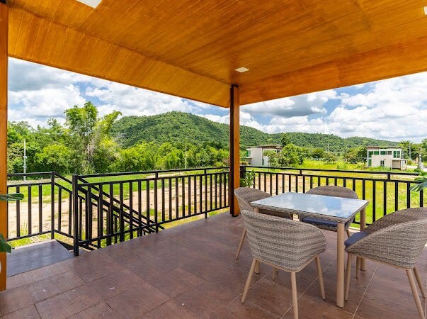 A covered outdoor patio features a glass-top table surrounded by four woven chairs, offering a relaxed dining space. Lush greenery and rolling hills are visible in the background, with a clear blue sky above.