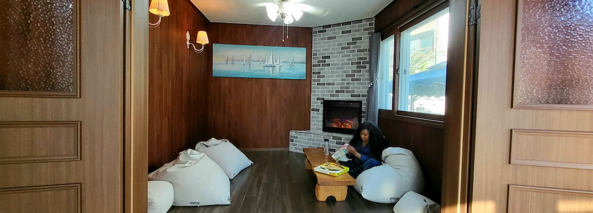 A cozy lounge area is presented, featuring warm wooden walls and a decorative stone fireplace. Soft, round cushions are arranged on the floor, while a wooden coffee table holds books. Natural light enters through large windows, complemented by ceiling lights.