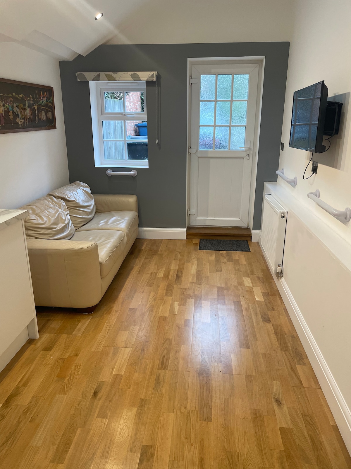 A cozy corner of the flat features a light-colored sofa with cushions, positioned against a wall that showcases artwork. Natural light enters through a small window beside the entrance door. The wooden floor adds warmth to the space, enhancing the inviting atmosphere.