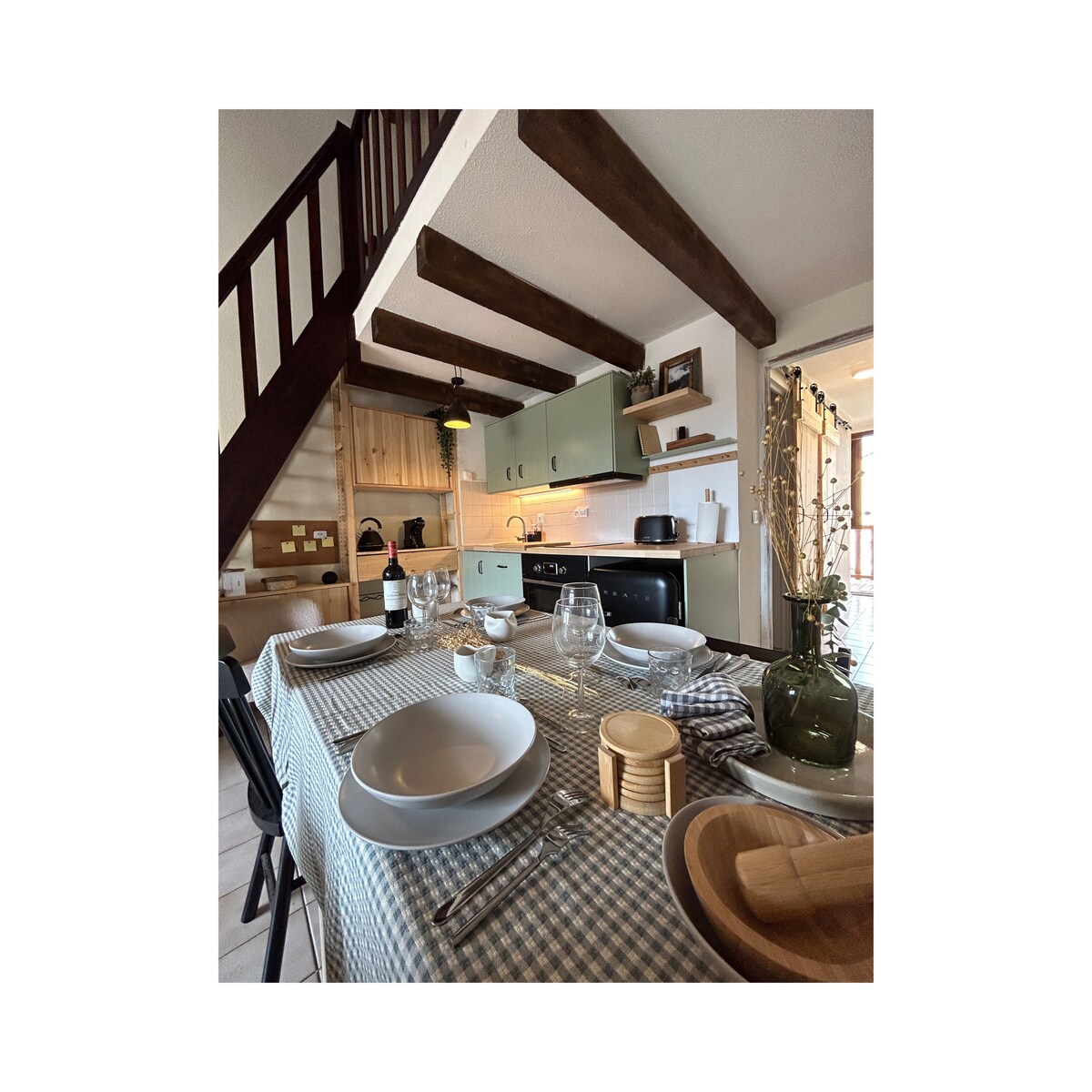 A dining table is set with plates, glasses, and utensils, surrounded by wooden chairs. The kitchen, featuring modern cabinetry and appliances, is visible in the background, with warm lighting highlighting the space. Exposed wooden beams add a rustic touch to the setting.
