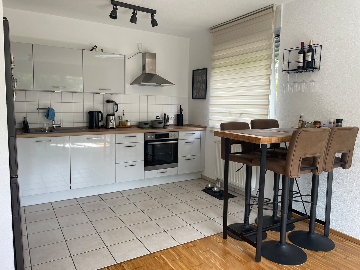 The kitchen features modern cabinetry and appliances, including an oven, stovetop, and a coffee maker. A high table with three stools provides a dining area. Natural light filters through large window shades, illuminating the tiled floor and the overall contemporary design.