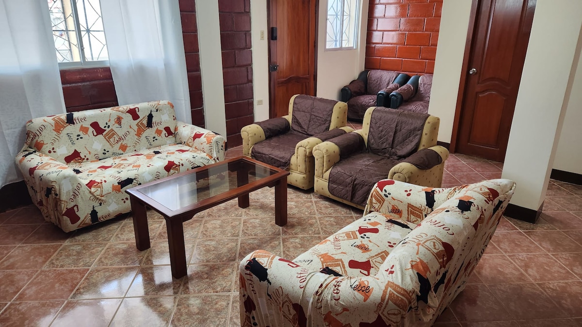 A living area features two patterned sofas and a coffee table, arranged around a cozy space. In the background, a sofa bed is visible, providing additional seating options. Light enters through the windows adorned with white curtains, enhancing the inviting atmosphere.