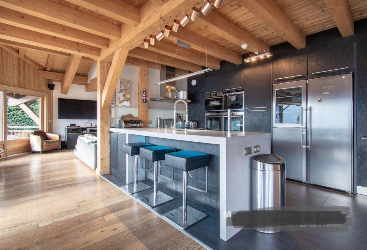 A modern kitchen is featured with a large island bar, complemented by high stools in a calming blue hue. Sleek appliances and cabinetry enhance the contemporary style, while natural wood beams provide warmth. An inviting open space connects to the living area, illuminating the room with natural light.