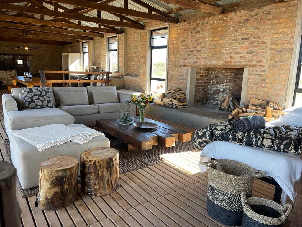 An open-concept living area features a cozy sectional sofa with decorative cushions, a rustic wooden coffee table, and two tree stump stools. A fireplace is situated in the background, surrounded by neatly stacked firewood. Natural light enters through large windows, illuminating the space.