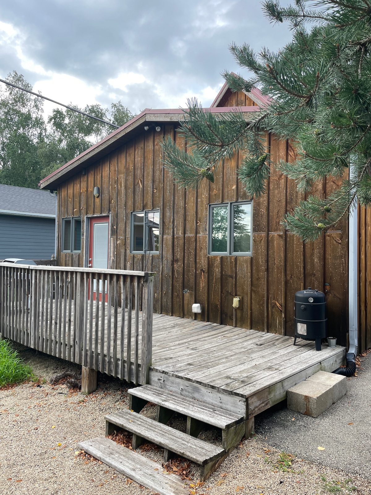 The exterior of a rustic wooden house features a sloped roof and a welcoming red door. A spacious deck, framed by railings, leads to a set of steps. Multiple windows allow natural light to enter, surrounded by greenery and gravel pathways.