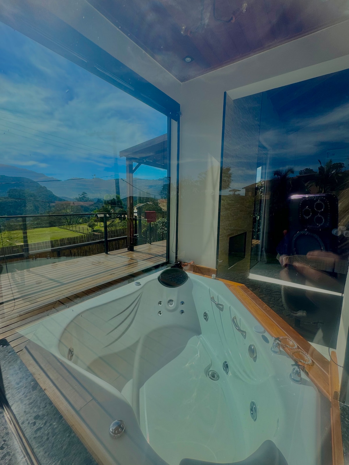 A spacious hot tub is positioned near large glass windows, allowing for scenic views of the surrounding mountains and outdoor deck area. Natural light enhances the relaxing ambiance, contributing to an inviting atmosphere perfect for unwinding.
