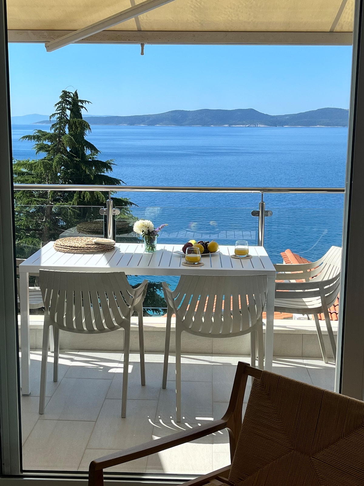A balcony is shown overlooking the ocean, featuring a white table set for two with fresh flowers and a few items. Light-colored chairs surround the table. The expansive sea view includes distant islands under a clear blue sky, creating a serene atmosphere.