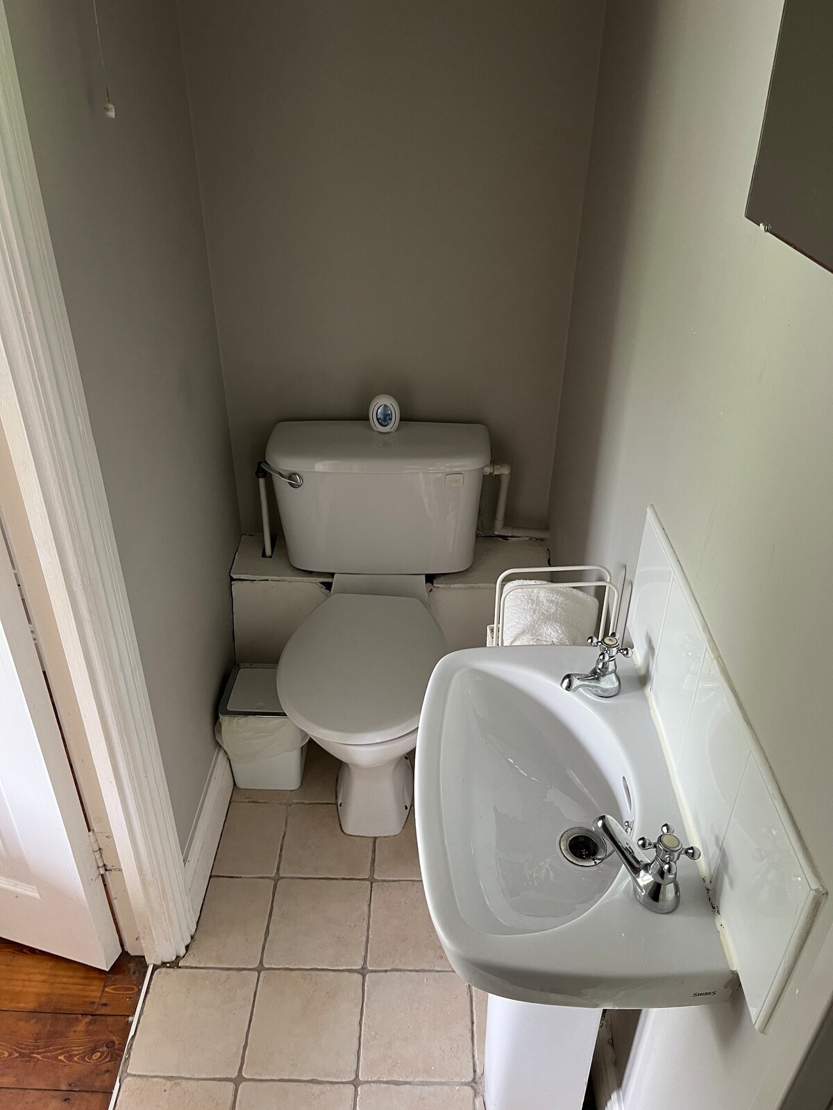 The compact bathroom is equipped with a white toilet and a small sink. Neutral-colored tiles cover the floor, and light grey walls provide a clean backdrop. An organizer for toiletries is positioned beside the sink, contributing to the functionality of the space.