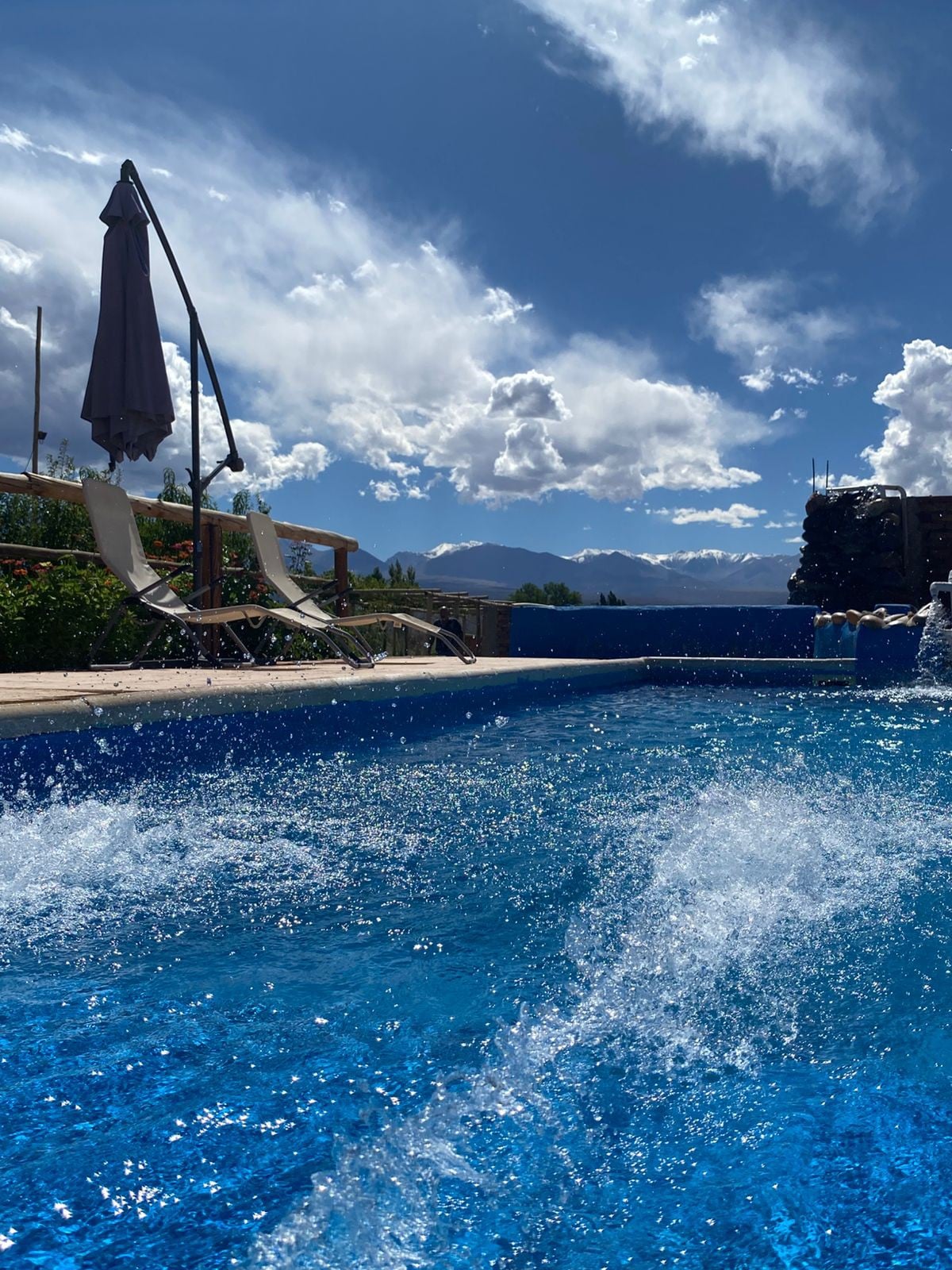 A swimming pool is situated amidst lush greenery, reflecting the clear blue sky. Lounge chairs are positioned nearby, accompanied by a parasol for shade. The stunning backdrop of the Andes mountains is visible, adding to the serene atmosphere.