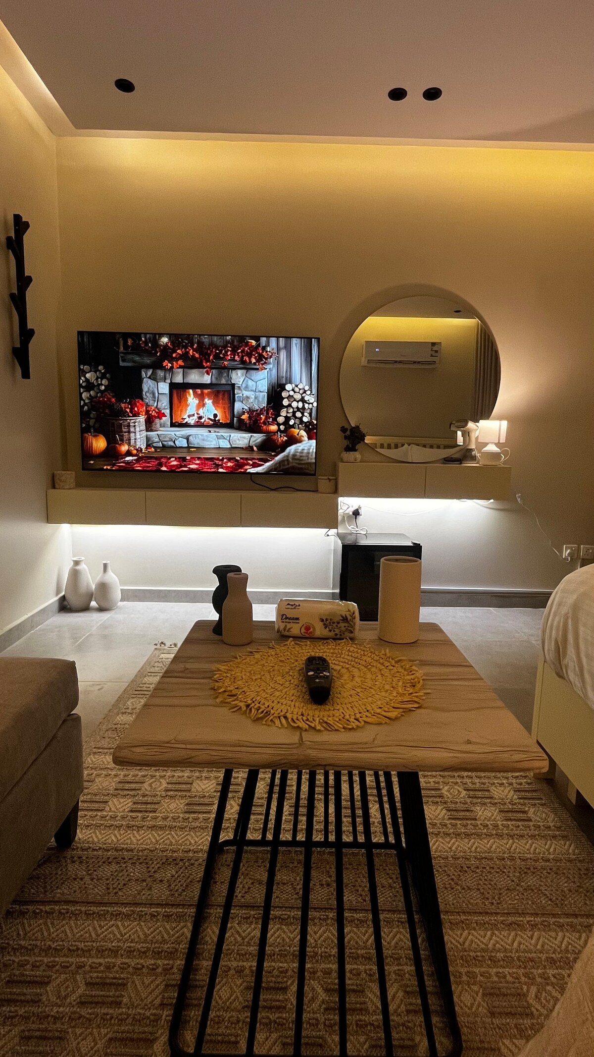 A cozy sitting area features a neutral-toned couch facing a modern television displaying a fireplace scene. A wooden coffee table with a decorative centerpiece is situated on a textured rug. Soft lighting enhances the warmth of the space, complemented by a mirror and minimalistic decor.