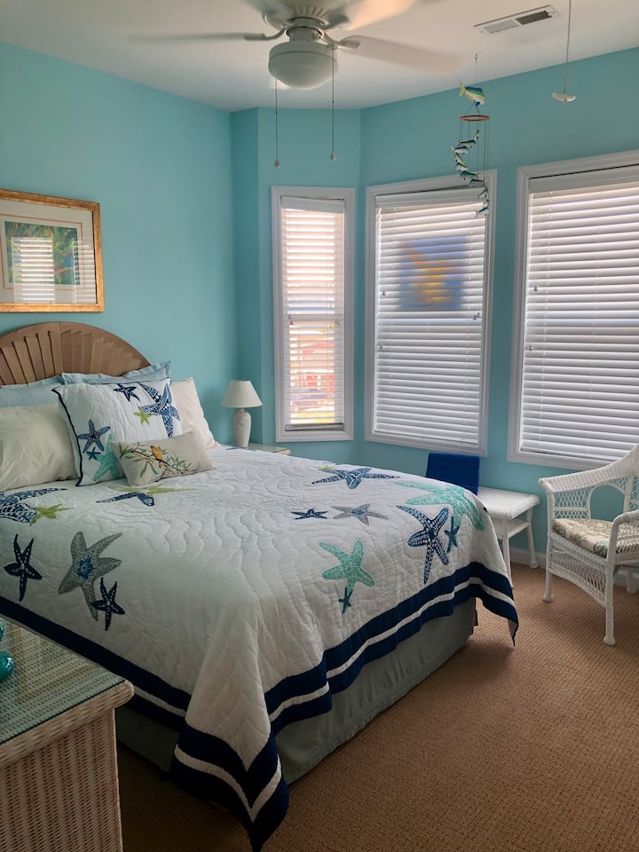 Dormitorio temático costero 3 en el tercer nivel con una cama tamaño queen, 2 mesitas de noche y un tocador de tamaño completo, un banco de equipaje y un sillón de mimbre. 