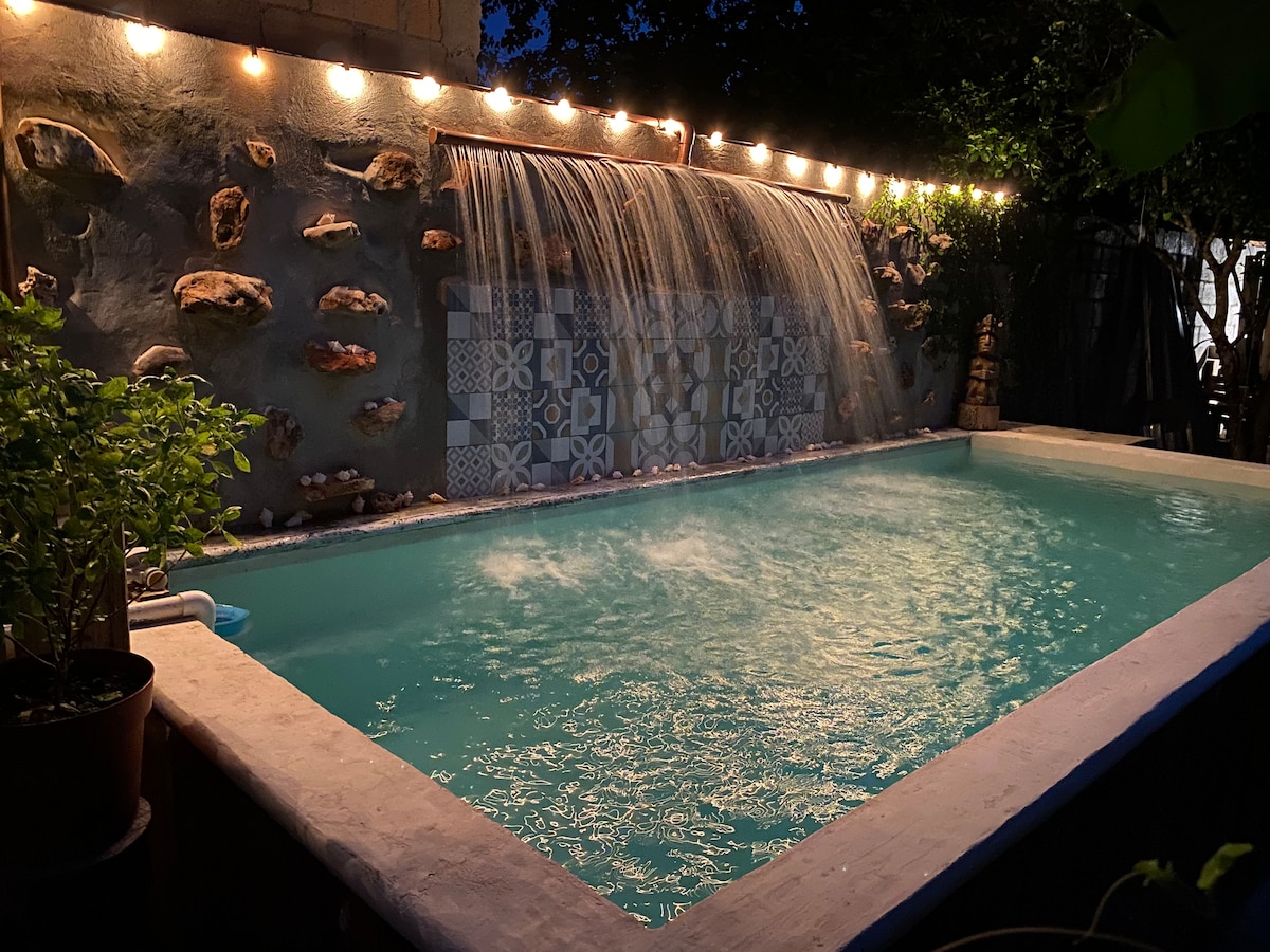 A tranquil pool area is illuminated by soft, warm lights strung overhead. A gentle waterfall cascades down a stone wall, blending with the calm water of the pool. Surrounding greenery adds a natural element, enhancing the serene ambiance of the outdoor space.