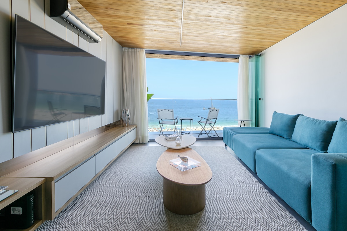 A living area features a comfortable sectional sofa in turquoise, a round coffee table, and a large flat-screen TV mounted on the wall. Floor-to-ceiling glass doors reveal a breathtaking ocean view, with two outdoor chairs positioned on the balcony.