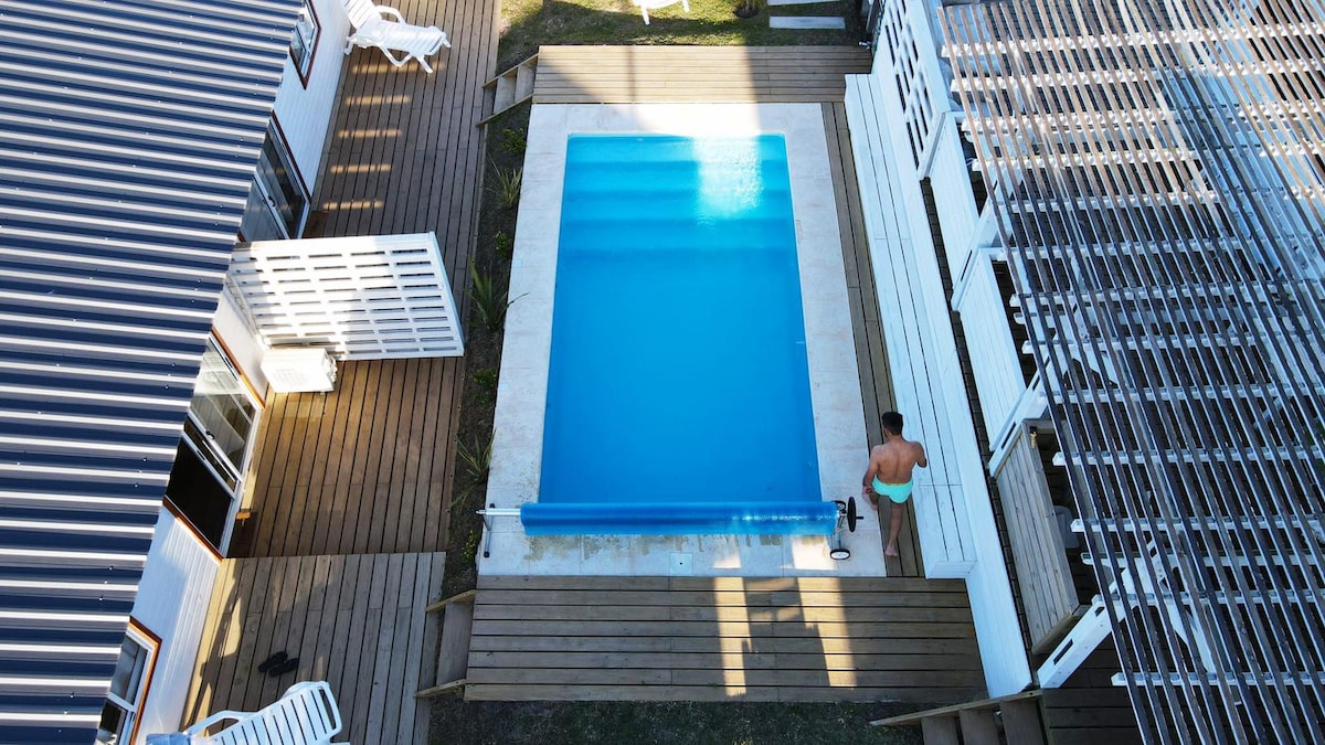 An aerial view captures a rectangular swimming pool surrounded by wooden decking. Two white lounge chairs are placed nearby, with a figure standing at the edge of the pool. The layout includes buildings with varied roofing, emphasizing the outdoor space's openness.