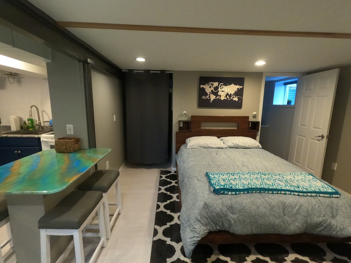 A modern studio space features a spacious queen bed with a patterned blanket and decorative throw. A compact kitchen area is visible with blue cabinetry. A small dining table with stools is present, along with a large area rug adding texture to the room.