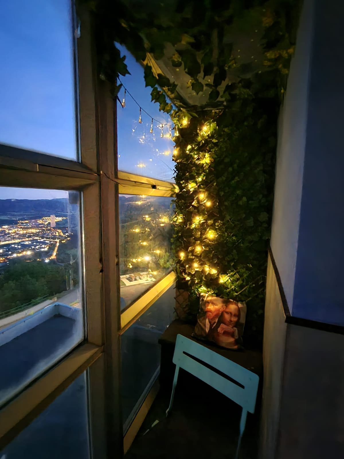 A cozy corner features a small table and a light blue chair, surrounded by lush greenery and illuminated by warm fairy lights. Large windows offer views of Jaca, enhancing the inviting ambiance of the space as evening falls.