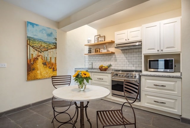 A bright kitchen area features white cabinetry and modern appliances, including a microwave and stove. A round marble table is surrounded by four stylish metal chairs. A colorful painting of a village scene adds a touch of artistry to the space.