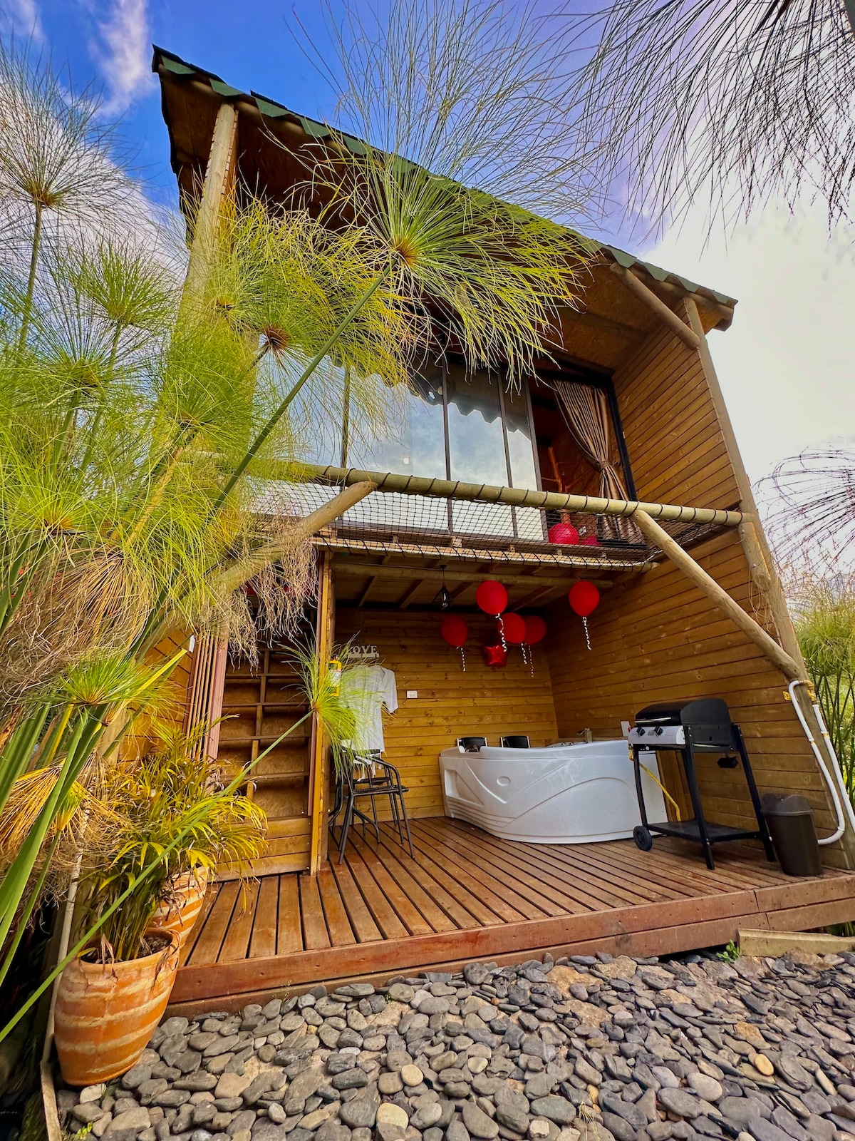 A wooden cabin is surrounded by lush greenery, with a private outdoor jacuzzi visible on the deck. The entrance features a small staircase leading to large glass doors, allowing natural light to brighten the space. Decorative lanterns add a welcoming touch.
