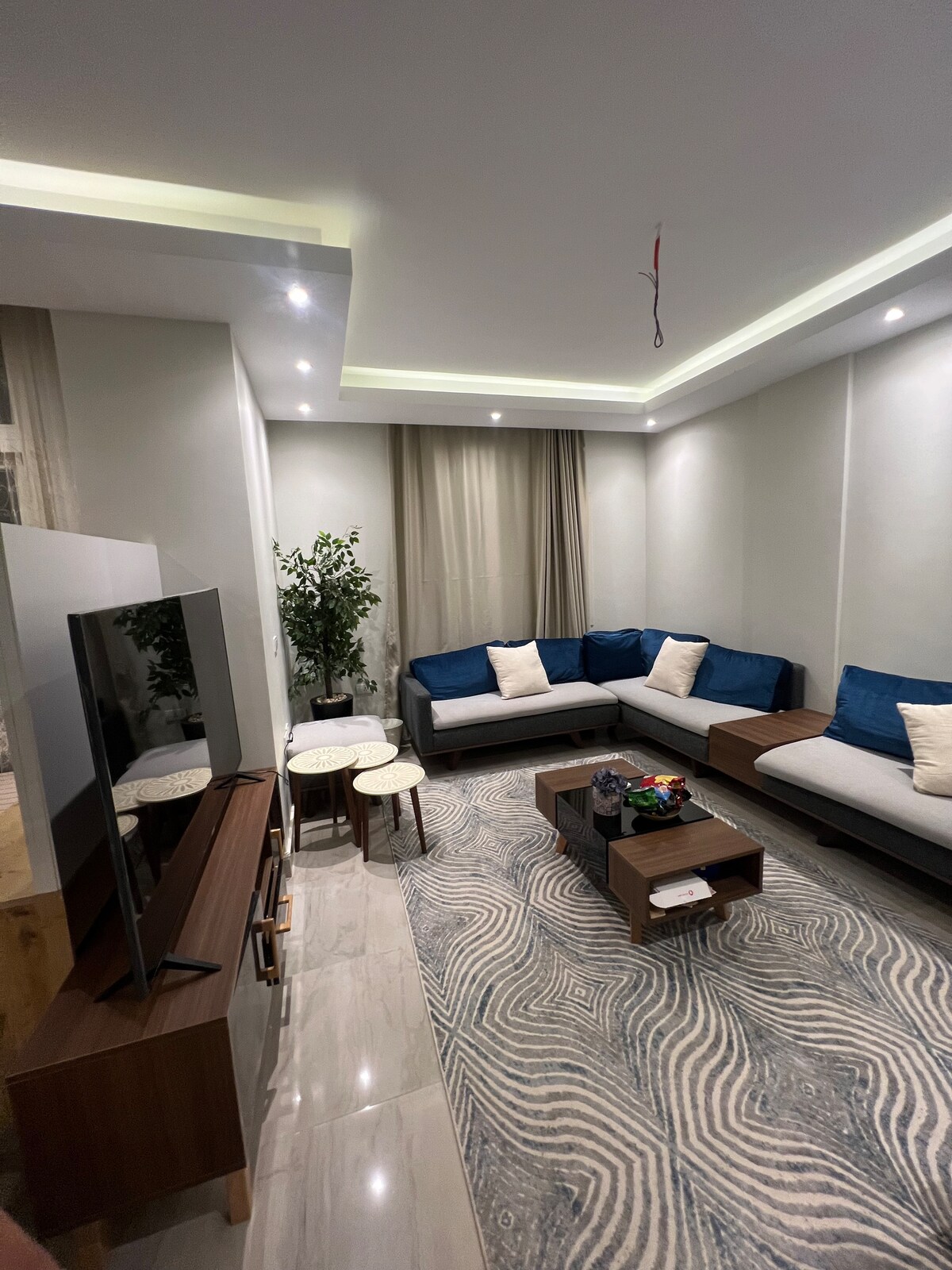 A modern living room features a comfortable sectional sofa with blue accents and neutral cushions. Two stylish side tables are positioned next to a wooden TV stand, which holds a flat-screen television. Soft lighting enhances the space, complemented by a decorative area rug that adds warmth.
