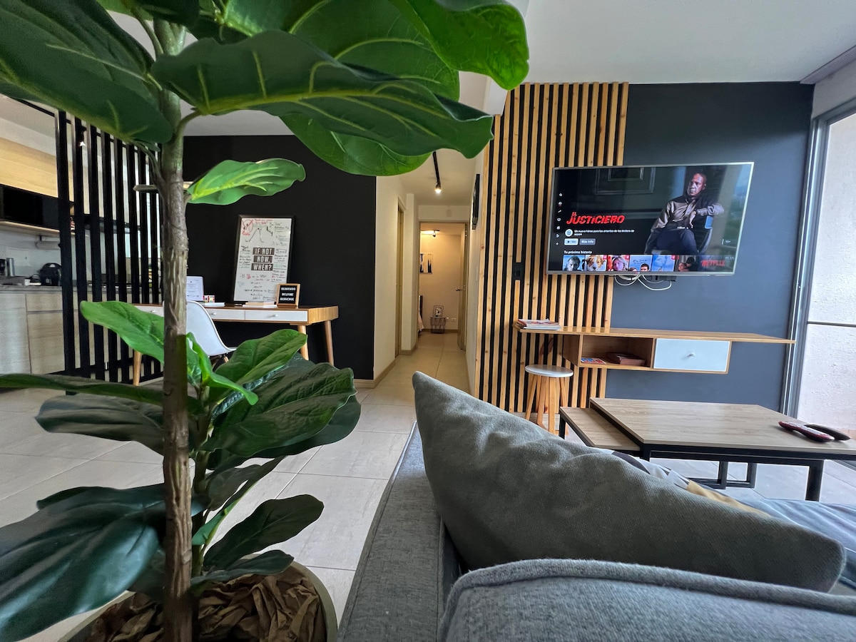 A modern living area features a comfortable gray sofa facing a wall-mounted television. A decorative plant adds greenery to the space. In the background, a dining table and kitchen area are visible, with warm wooden accents enhancing the contemporary design.