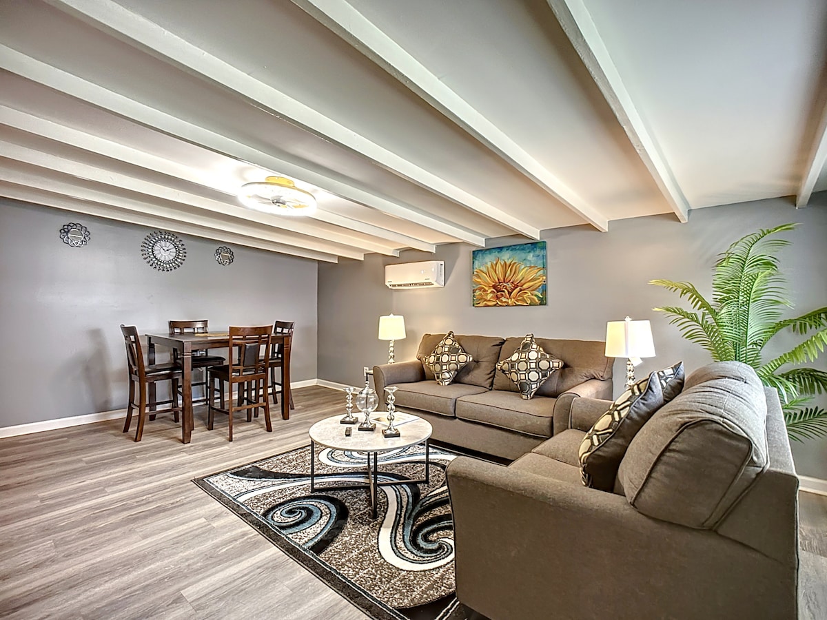 The living area features a comfortable seating arrangement with gray sofas and decorative pillows, complemented by a round coffee table. A dining area with four chairs is visible against a backdrop of modern decor, and split-type air conditioning is positioned overhead. Beam ceilings add to the spacious feel.