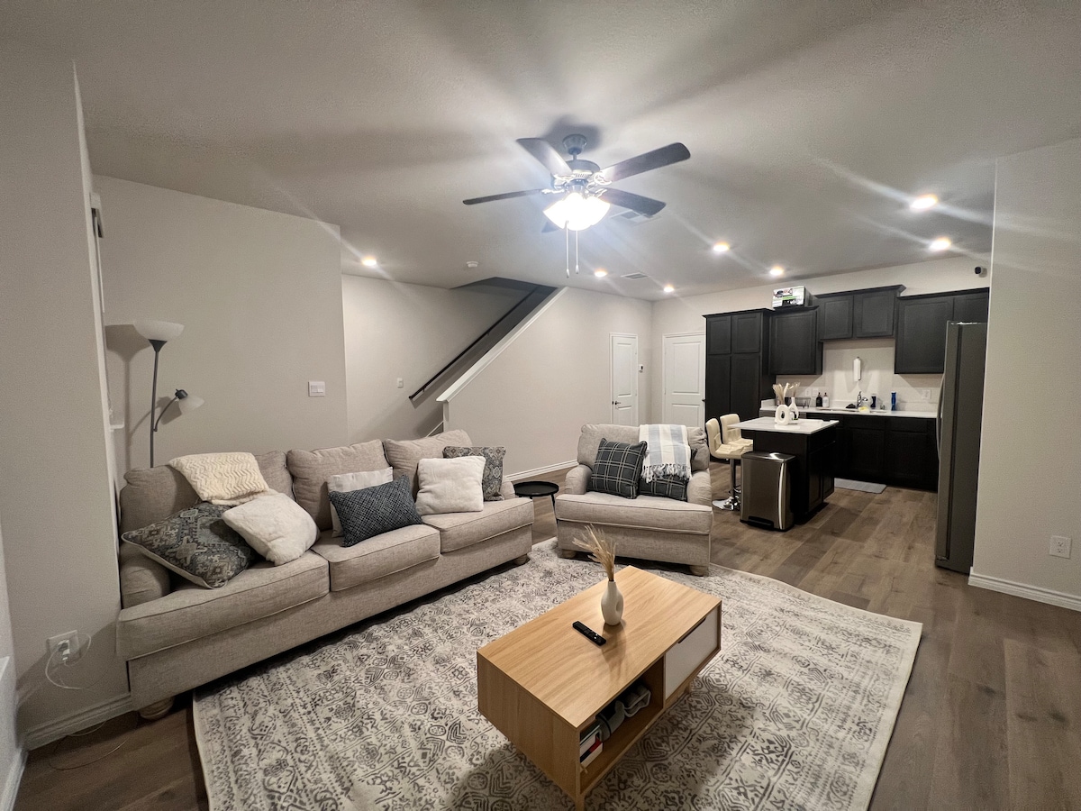 A spacious living area features a neutral-toned sectional sofa paired with decorative pillows, centered around a low wooden coffee table. Light-colored hardwood flooring complements the soft area rug, while a ceiling fan and recessed lighting enhance the comfortable ambiance. A dining area and kitchen are visible in the background.