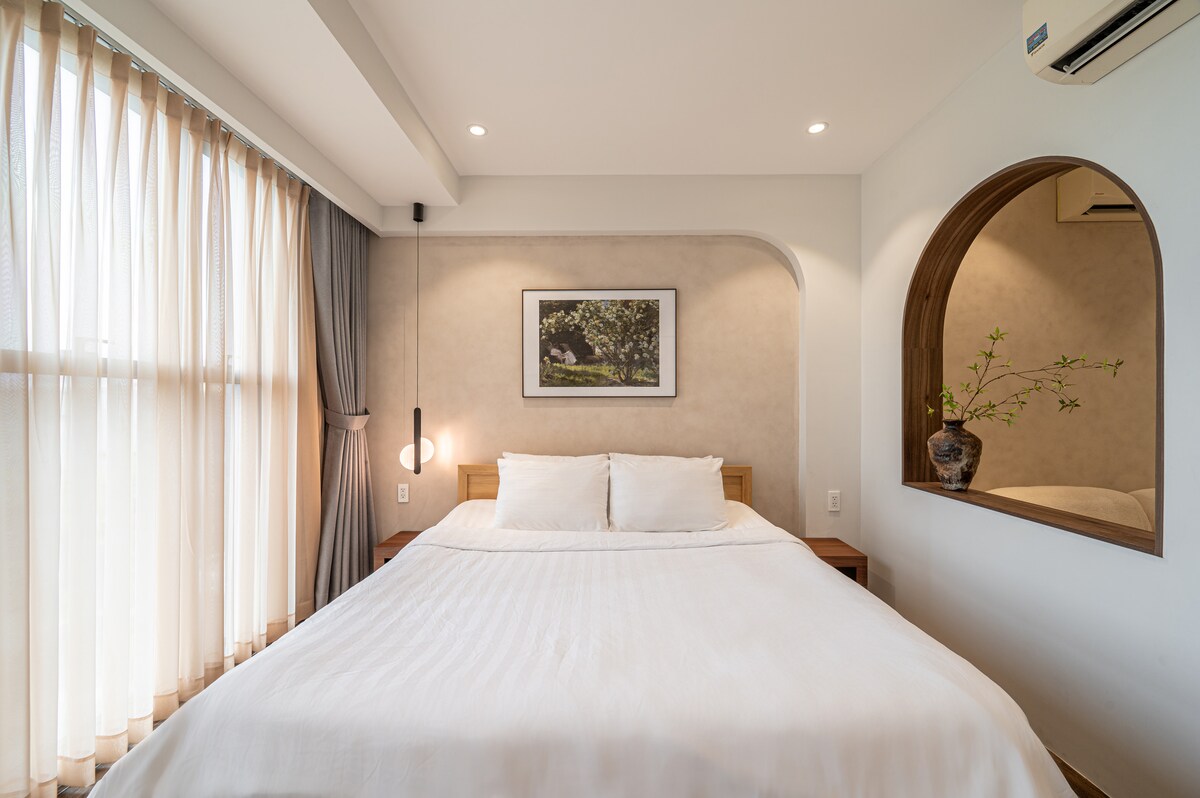 The spacious sleeping area features a large bed with neatly arranged white linens. Natural light fills the room through floor-to-ceiling curtains. A decorative mirror is seen on the wall, with a photo depicting a landscape positioned above the bed, complemented by a vase on the nightstand.