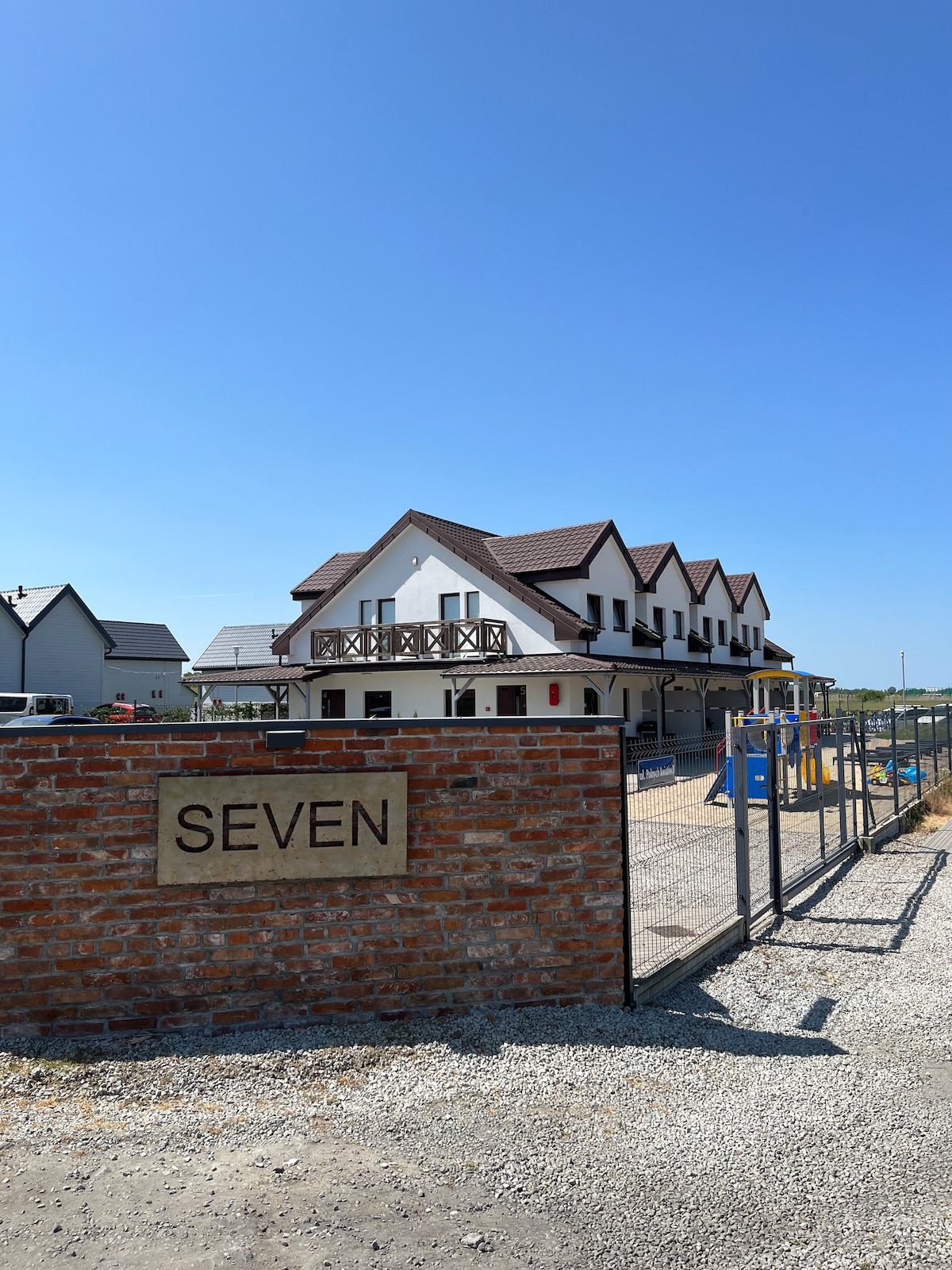 The entrance to the property features a stone wall with a sign reading 'SEVEN.' The building is a multi-story structure with a pitched roof, balconies, and a bright blue sky overhead. A well-maintained pathway leads up to the entrance.