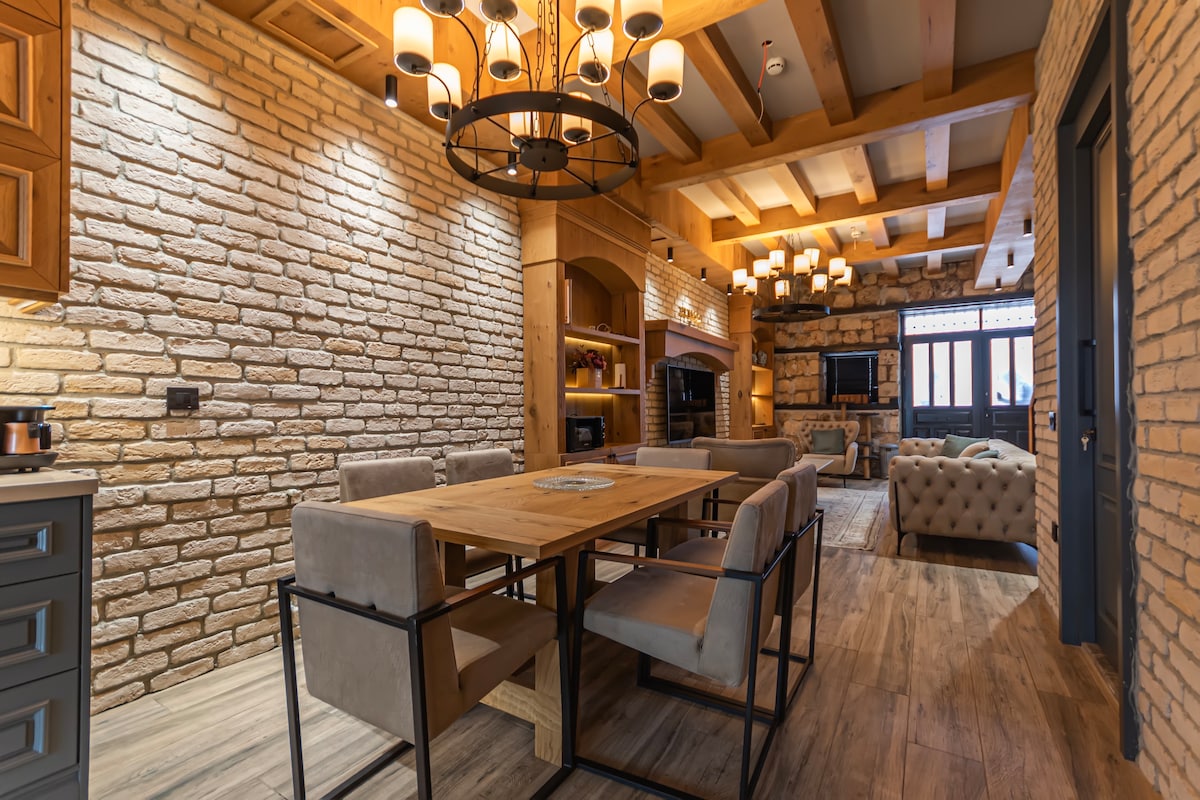 A dining area features a wooden table surrounded by several upholstered chairs. Exposed brick walls and wooden beams add character to the space. Soft lighting is provided by multiple hanging fixtures, highlighting the warm tones of the decor and creating a welcoming environment.