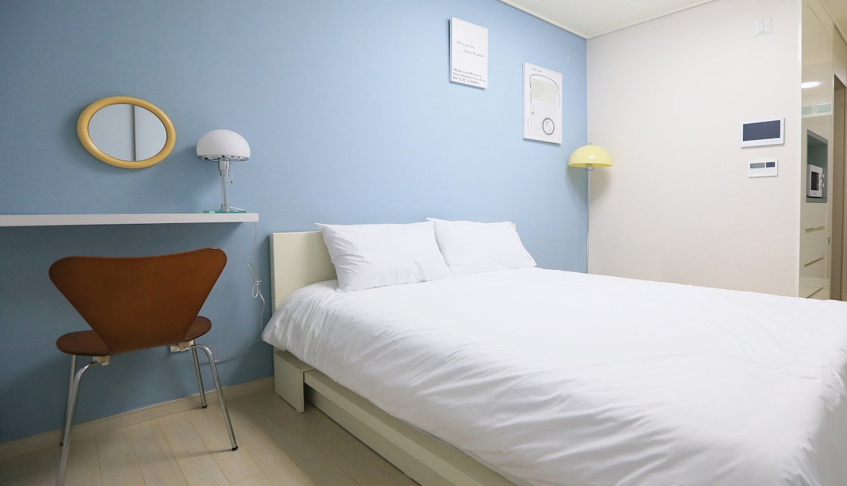 A bright sleeping area features a spacious bed with crisp white linens against a soft blue wall. A simple desk and a stylish chair are positioned nearby, along with a lamp that provides a warm glow, creating a functional yet inviting space.
