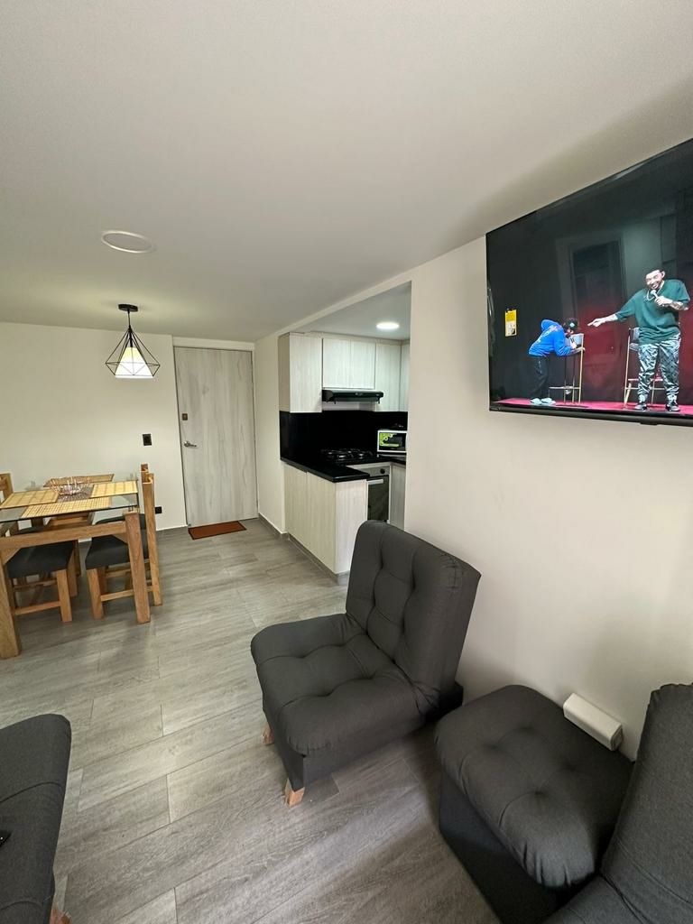 A cozy living area features two dark upholstered chairs facing a wall-mounted television. A wooden dining table with chairs is positioned nearby, alongside a compact kitchen area with modern cabinetry and appliances. Natural light illuminates the space through a ceiling fixture.