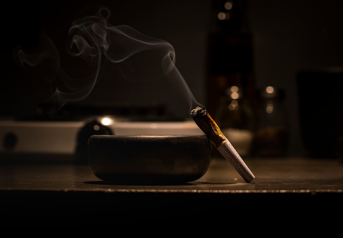 A close-up of a lit cigarette resting on a dark ashtray, with wisps of smoke rising gracefully into the air. The background features faint outlines of kitchen items, suggesting a warm and intimate setting.