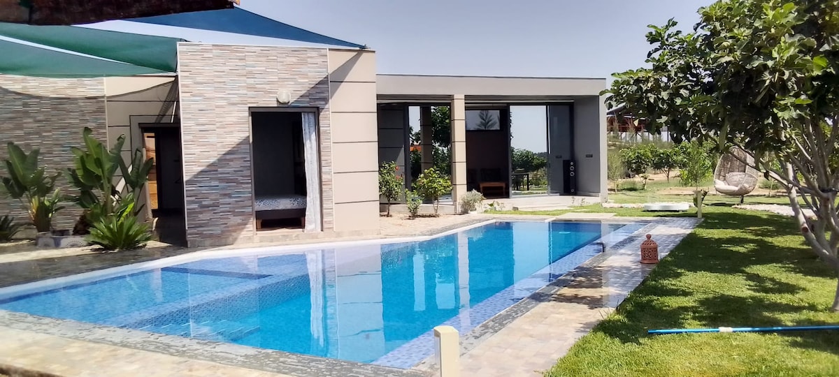 A private pool is framed by a well-maintained garden featuring lush greenery and trees. Adjacent to the pool, a modern structure with large windows offers views of the outdoor space. The area is designed for relaxation, highlighted by a comfortable seating arrangement near the pool.